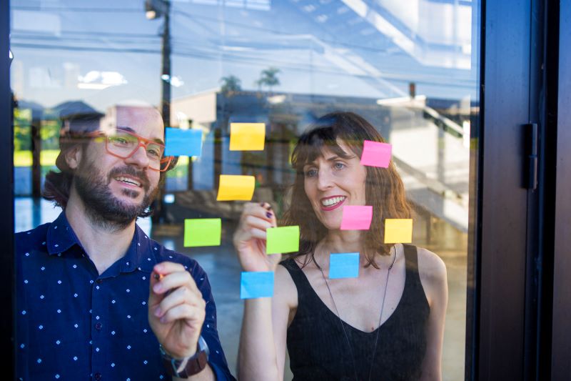 mapa de stakeholders: homem e mulher em escritório analisando post-its em painel de vidro